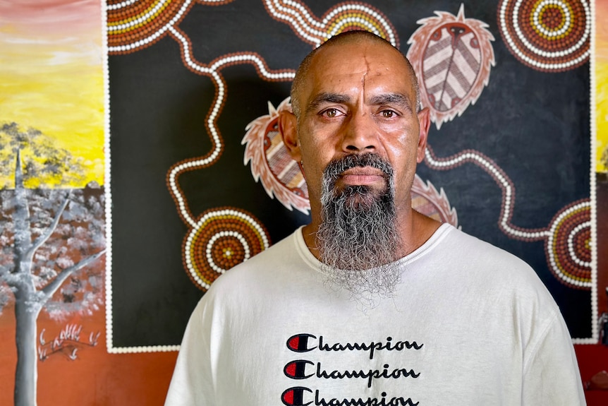 Bearded Aboriginal man wearing a white champion T-shirt stands in front of traditional artwork.