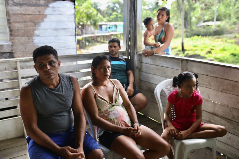 Residents gathered around the wooden deck area, some holding small children. The greenery of the Amazon rainforest can be seen outside the deck.