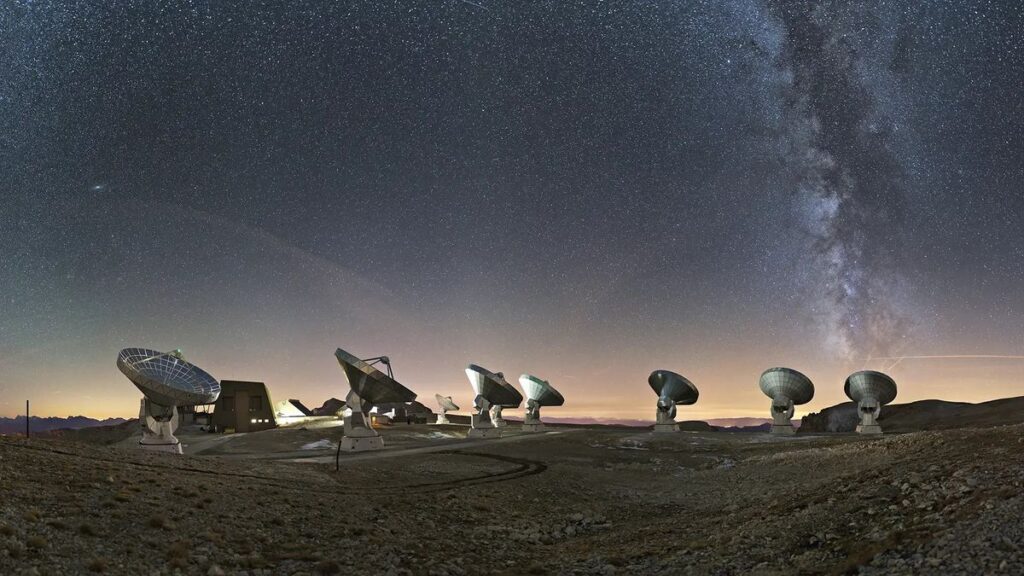 several giant satellite dishes stand scattered in a vast rolling field, pointed toward the night sky and an array of stars next to the arm of the milky way