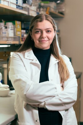 A woman leans on a lab bench, crosses her arms, and smiles at the camera. She was wearing a black shirt, white lab coat and gold earrings. She has light skin and chest-length brown hair with blonde highlights that is slicked back from the front. 