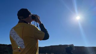 A man in a yellow T-shirt looks at the sun with binoculars.