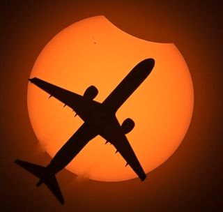 The silhouette of an airplane passes in front of the solar disk, which is missing a patch of moon-covered space in its upper right corner.