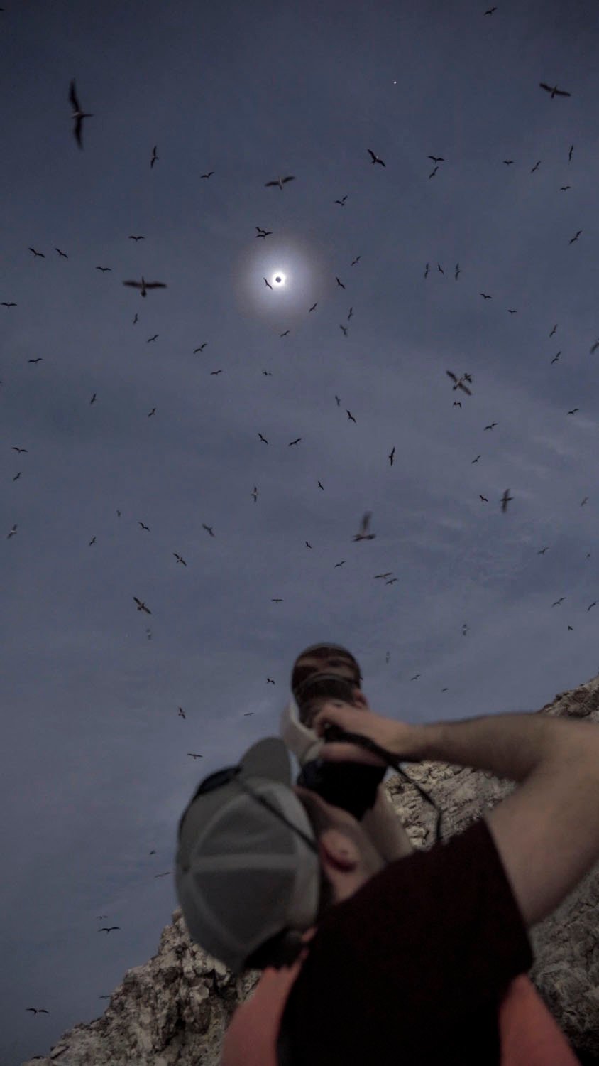 Under a dim sky, a man captures a lunar eclipse with a camera, surrounded by countless flying birds.