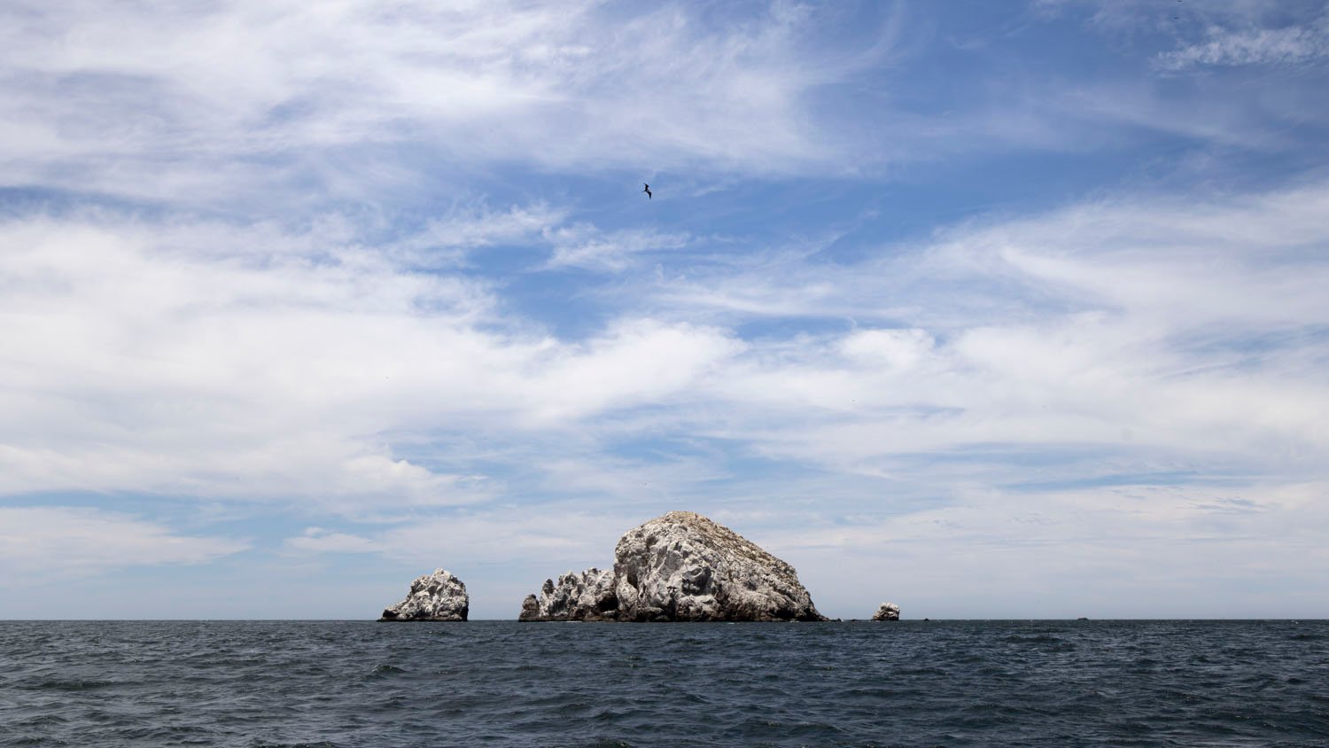 Under a cloudy sky, a large craggy rock formation is surrounded by a slightly choppy sea, and a bird flies high.