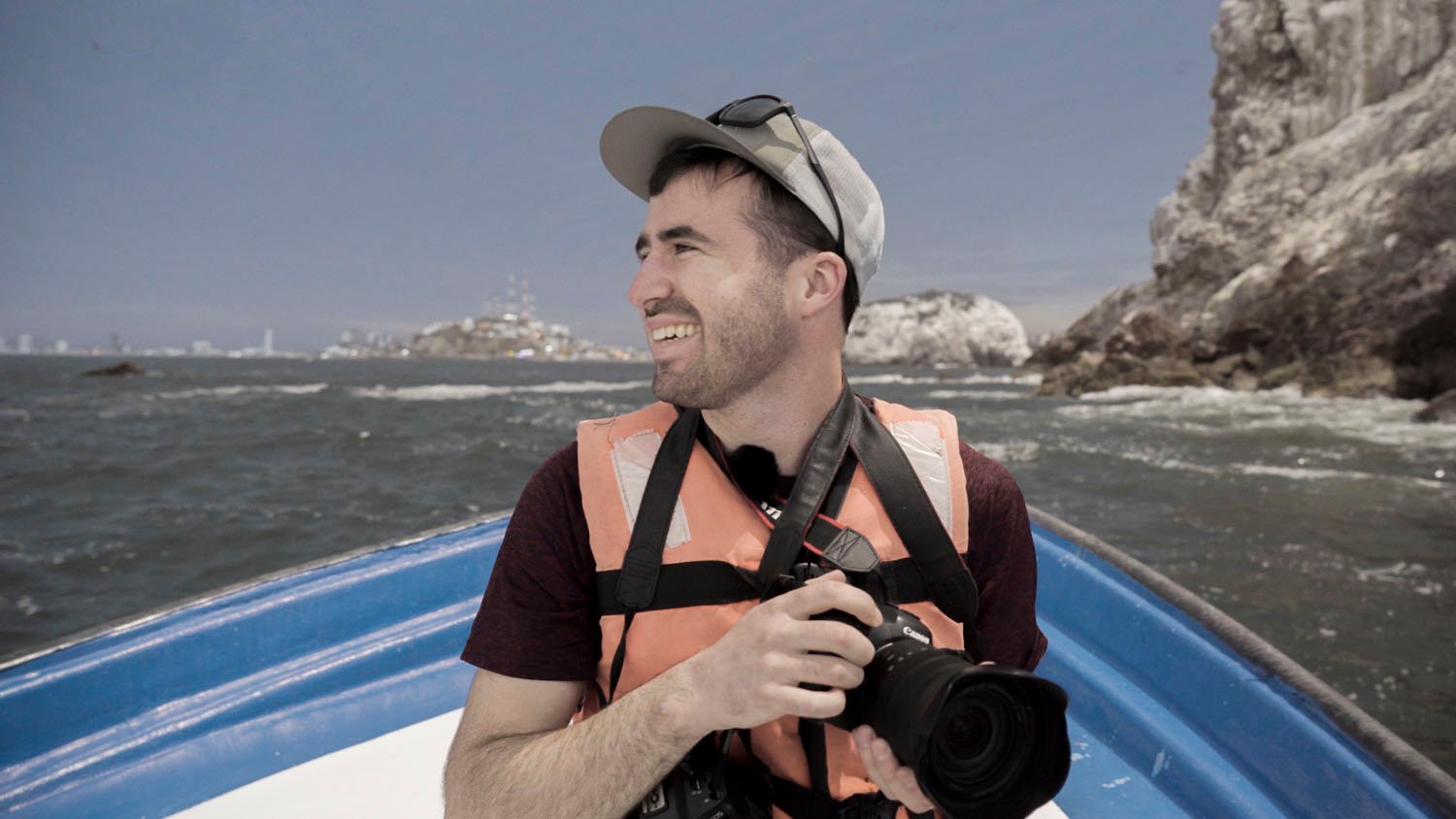 A smiling man wearing a life jacket and hat sits on a boat holding a camera with a rocky shoreline and water in the background.
