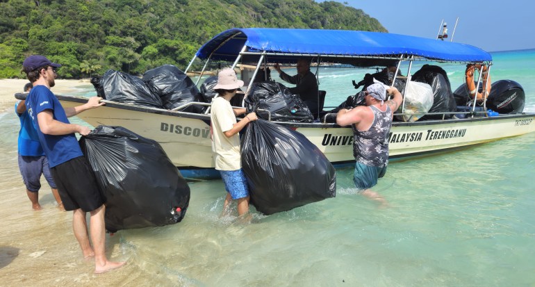 A group of men in shorts and T-shirts loaded large garbage bags into a small boat near the beach. The water is clear. 