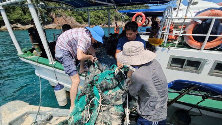 A group of men moved a large pile of discarded fishing nets onto a small boat. The net is green and gray and knotted. 