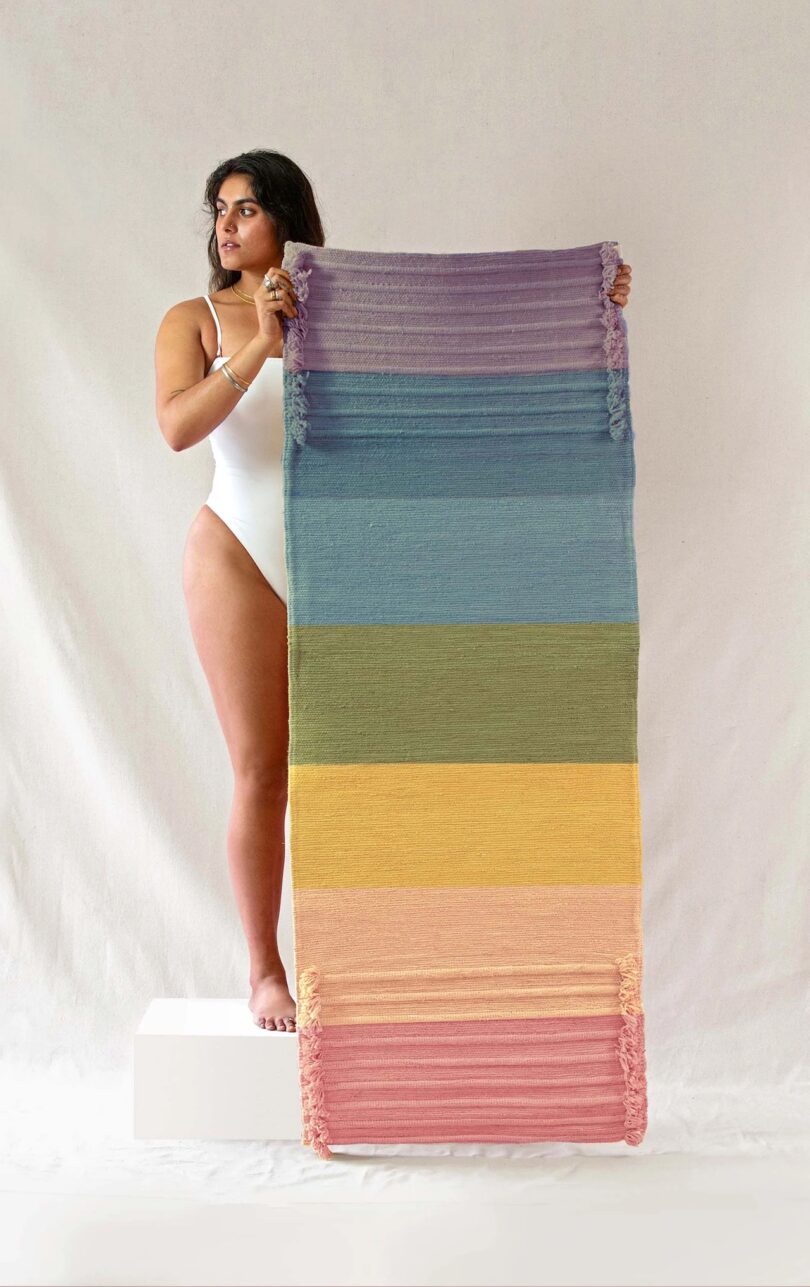 A woman holds an Oko Living Chakra Energy - Herbal Yoga Mat; from top to bottom, the mats are purple, blue, light blue, green, yellow, peach/light orange, and dark pink