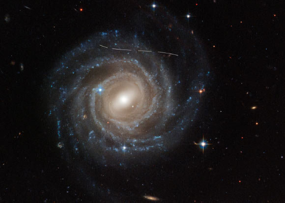 This Hubble image of the barred spiral galaxy UGC 12158 looks like someone took a white marking pen to it. In reality it is a combination of time exposures of a foreground asteroid moving through Hubbles field-of-view, photobombing the observation of the galaxy. Several exposures of the galaxy were taken, what is evidence in the dashed pattern. The asteroid appears as a curved trail due to parallax: because Hubble is not stationary, but orbiting Earth, and this gives the illusion that the faint asteroid is swimming along a curved trajectory. The uncharted asteroid is in inside the asteroid belt in our Solar System, and hence is 10 trillion times closer to Hubble than the background galaxy. Image credit: NASA / ESA / Hubble / Pablo Garca Martn, UAM / Joseph DePasquale, STScI / Alex Filippenko, UC Berkeley.