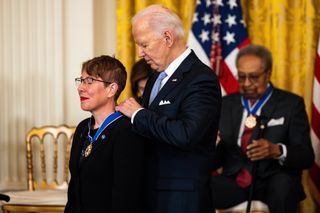 A woman wearing a NASA pin wears red lipstick and smiles, while an older man in a suit behind her pins the medal around her neck.