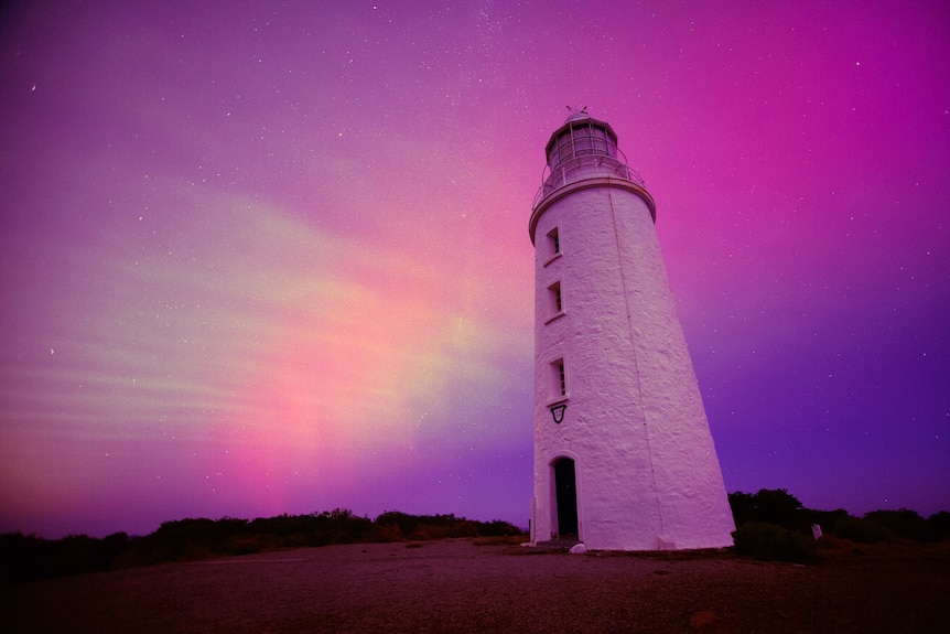 A lighthouse stands in front of a pink and purple sky illuminated by aurora