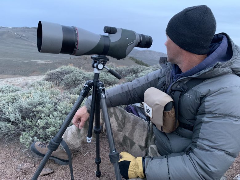 Nate Seward, dressed warmly, sat on the ground in a wilderness area with a large pair of binoculars on a tripod.